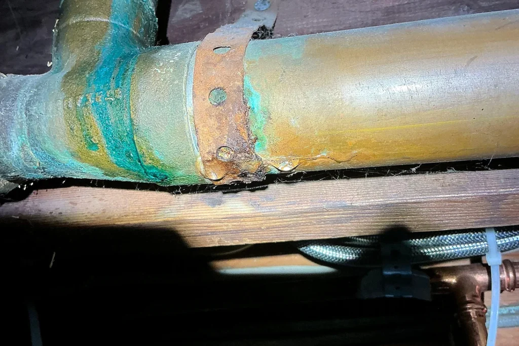Close-up of a badly corroded pipe with green verdigris and rust, plus a rusted-through hanger strap, before Empire State Plumbing replaced it