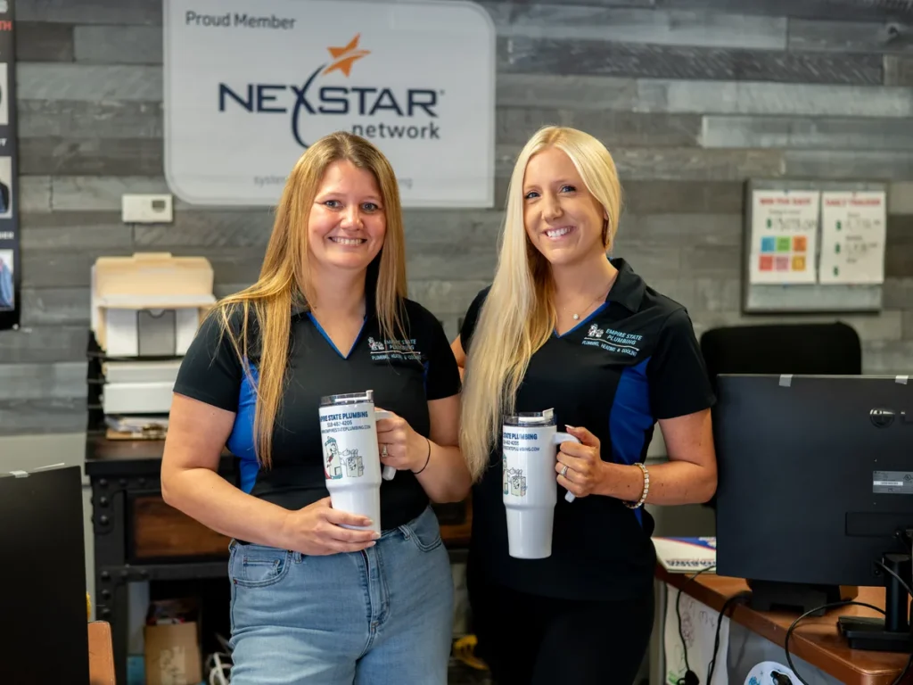 Two Empire State Plumbing office team members holding ESP travel mugs beneath the Nexstar Network Proud Member sign at the Albany office